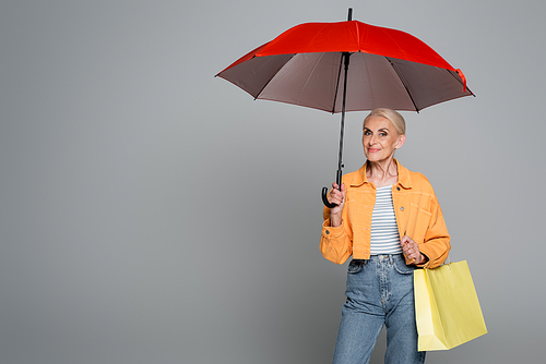 stylish senior woman with shopping bag  under red umbrella on grey