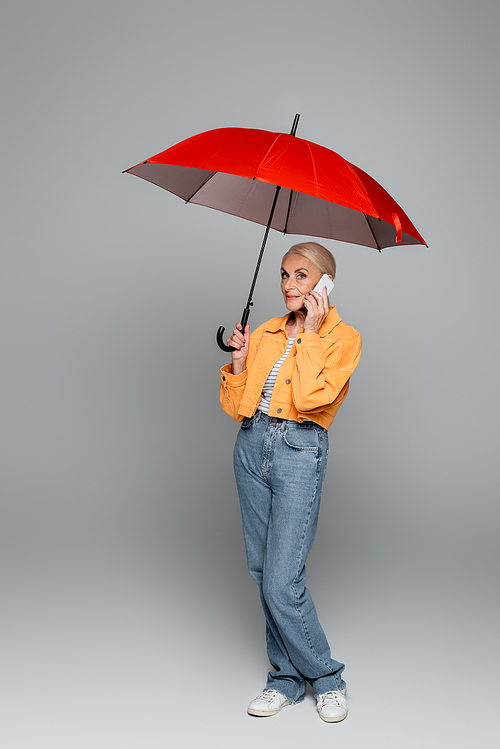 elderly woman in jeans and orange jacket talking on cellphone under red umbrella on grey