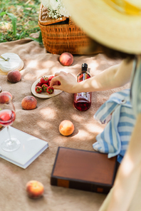 Blurred woman taking strawberry near wine and books on picnic blanket