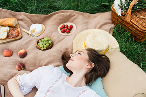 Top view of young woman lying near food and sun hat on blanket on grass