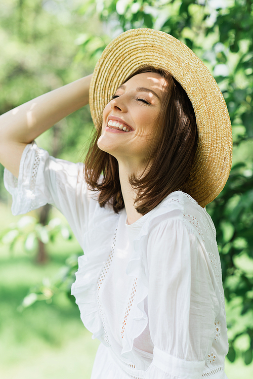 Happy woman in white dress holding sun hat in park