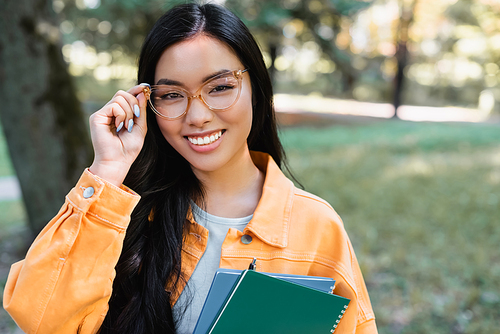 brunette asian student adjusting eyeglasses while holding notebooks in park
