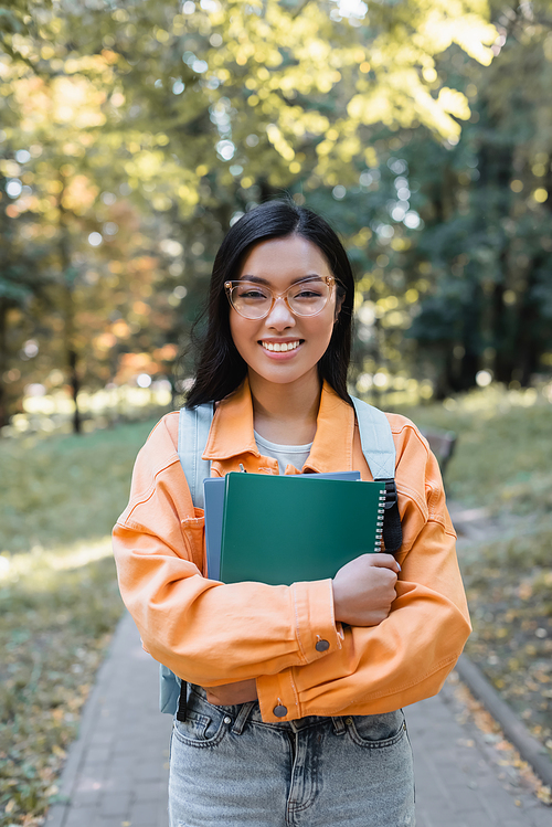 brunette asian student in eyeglasses smiling at camera while holding notebooks in park