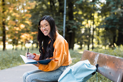 happy asian woman writing in notebook and smiling at camera on bench in park