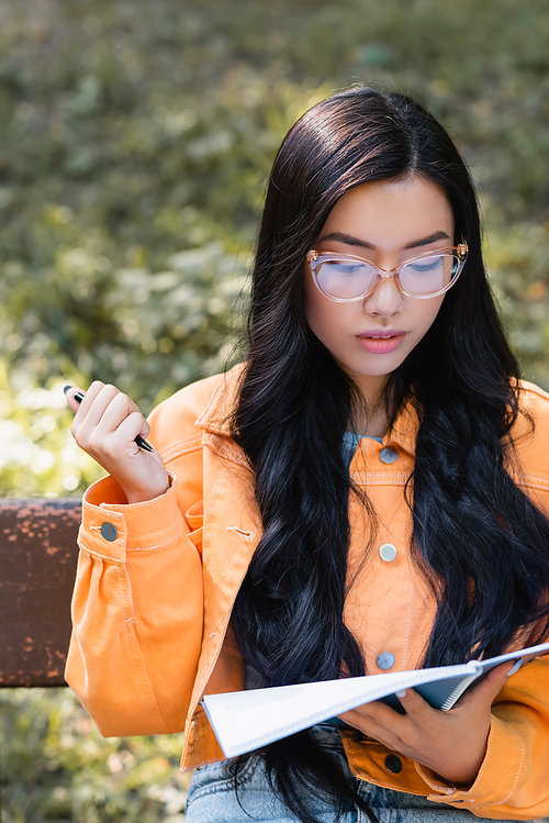 asian student in eyeglasses looking in notebook while sitting on bench in park
