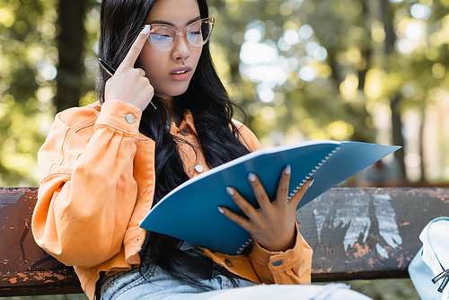 thoughtful asian student adjusting eyeglasses while looking in notebook in park