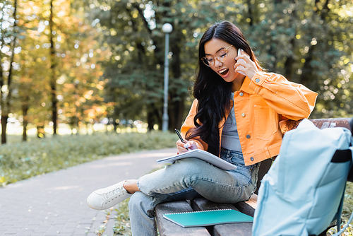 astonished asian woman writing in notebook while talking on smartphone on bench in park