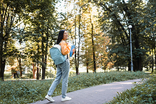 smiling asian woman in jeans walking in park with backpack and notebooks