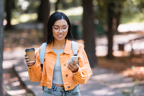 happy asian woman looking at mobile phone while holding takeaway drink outdoors