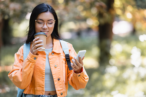 positive asian student drinking coffee to go while messaging on cellphone in park
