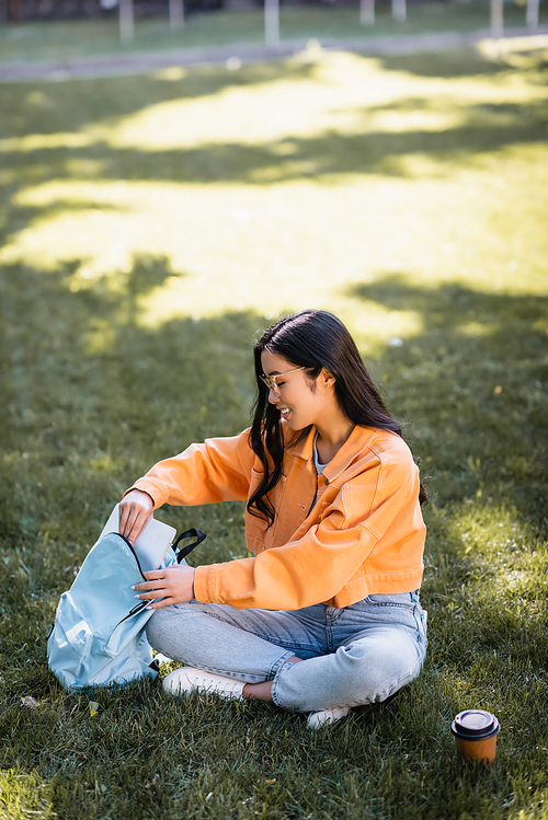 happy asian woman sitting on grass and putting laptop in backpack