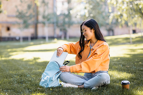 smiling asian woman putting laptop in backpack while sitting on grass with crossed legs