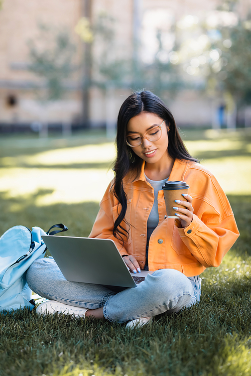 smiling asian woman using laptop while sitting on lawn with coffee to go
