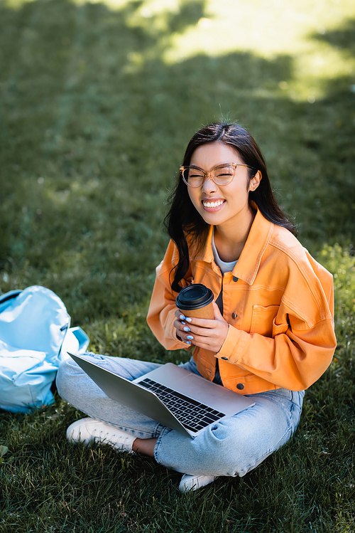 cheerful asian woman in eyeglasses sitting on grass with laptop and coffee to go
