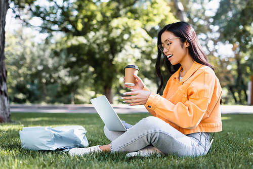 excited asian student pointing at laptop while sitting on lawn with coffee to go