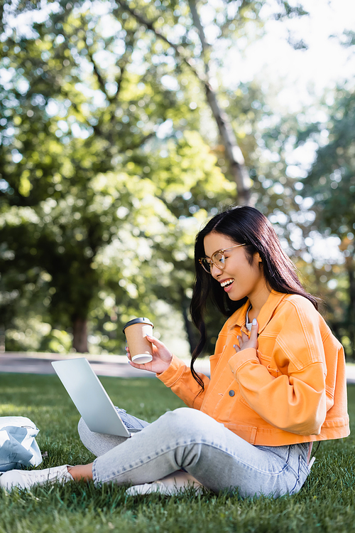 cheerful asian student with coffee to go touching chest while sitting on lawn with laptop