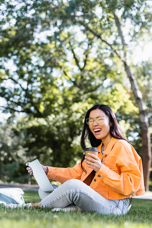 excited asian student with takeaway drink laughing while sitting on lawn with laptop