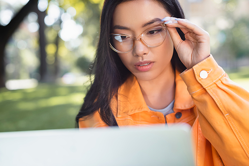 thoughtful asian woman touching eyeglasses while looking at blurred laptop outdoors