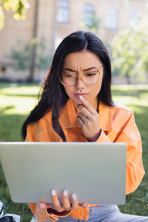 thoughtful asian woman in eyeglasses touching lips while looking at laptop in park