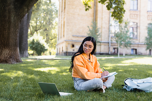 asian woman writing in notebook while sitting on lawn near laptop and backpack