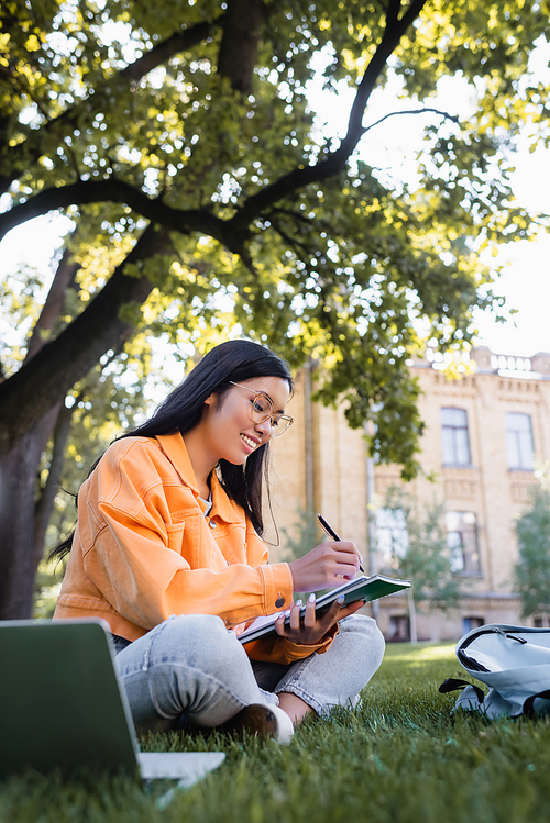 cheerful asian woman sitting in park and writing in notebook near laptop