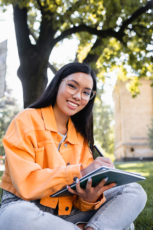 young asian woman smiling at camera while writing in notebook in park