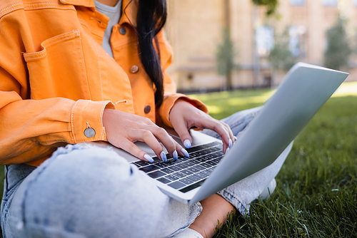 partial view of student in orange jacket sitting in park and typing on laptop