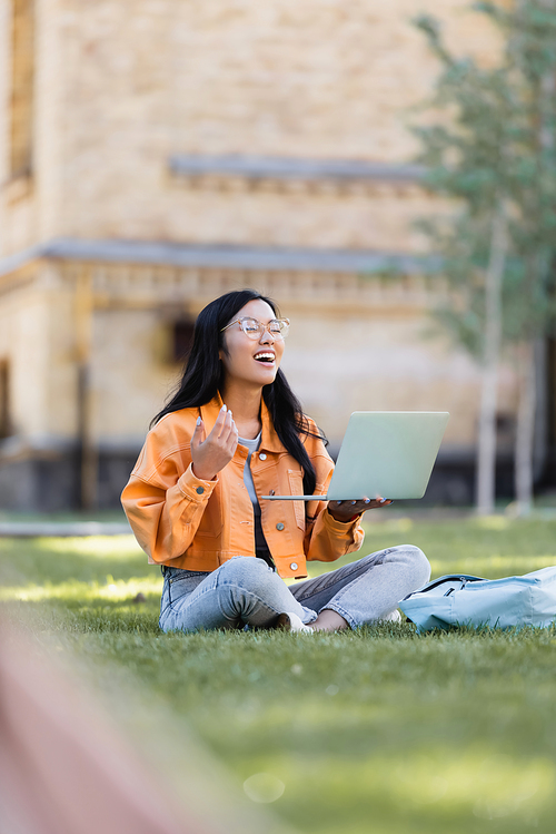 cheerful asian student laughing with closed eyes during video chat on lawn in park