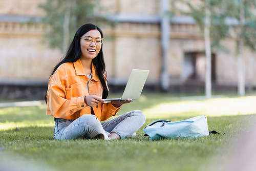 happy asian woman sitting on grass with laptop and smiling at camera