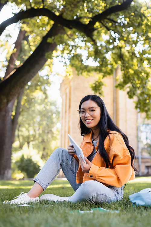happy asian student in eyeglasses  while sitting on lawn with digital tablet