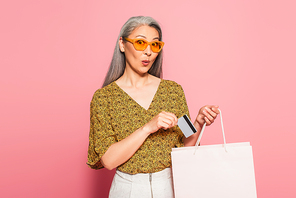 amazed middle aged woman putting credit card into shopping bag on pink background