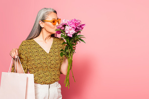 mature woman with shopping bag enjoying aroma of fresh peonies on pink background