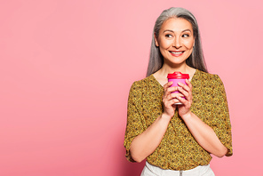 cheerful middle aged woman with coffee to go looking aside on pink background