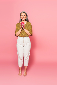 full length view of fashionable middle aged woman with paper cup on pink background