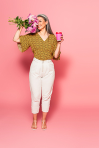 happy mature woman with takeaway drink obscuring face with peonies on pink background