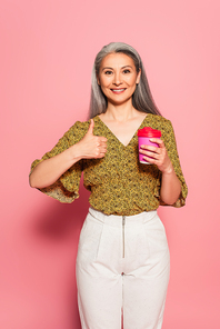 pleased woman with coffee to go showing thumb up on pink background