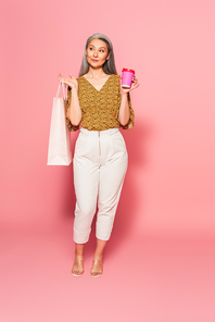 full length view of middle aged woman with paper cup and shopping bag looking away on pink