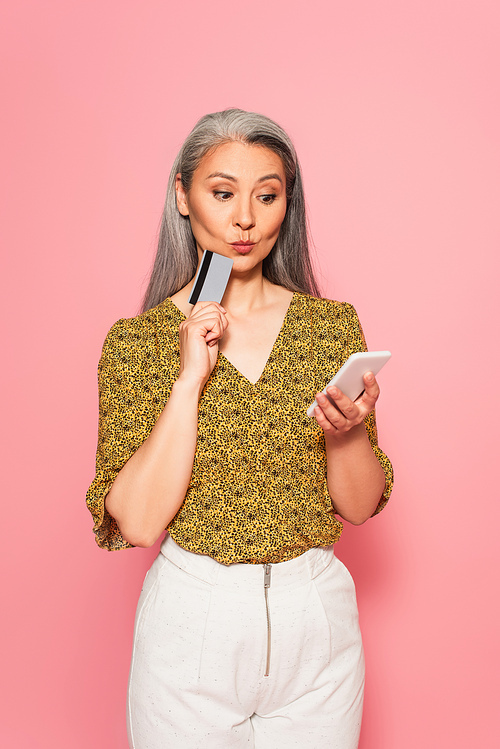 thoughtful mature woman holding credit card and cellphone isolated on pink