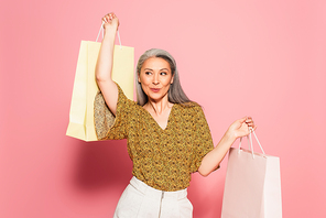 cheerful middle aged woman in stylish blouse posing with shopping bags on pink background