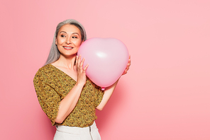 happy middle aged woman in patterned blouse holding heart-shaped balloon on pink background