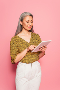 trendy middle aged woman using digital tablet while standing on pink background