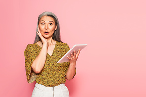 astonished woman with digital tablet touching face and  isolated on pink