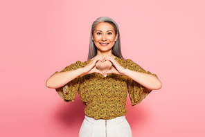 pleased woman in yellow blouse with pattern showing heart symbol on pink background