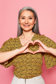 happy mature woman with grey hair showing heart sign with hands isolated on pink