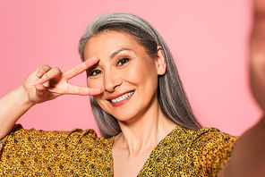 middle aged woman with grey hair showing victory sign near face while smiling isolated on pink