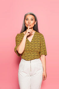 excited mature woman showing hush gesture while  isolated on pink