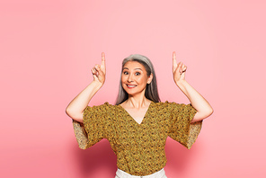 cheerful middle aged woman pointing up with fingers on pink background