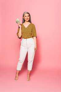 full length view of mature woman in white pants and blouse standing with dollar banknotes on pink