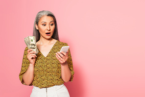 surprised middle aged woman with dollar banknotes looking at smartphone on pink background