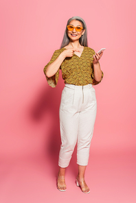 mature woman in stylish clothes and sunglasses pointing at smartphone on pink background